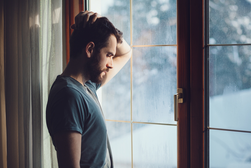 Man standing alone at window feeling defeated.